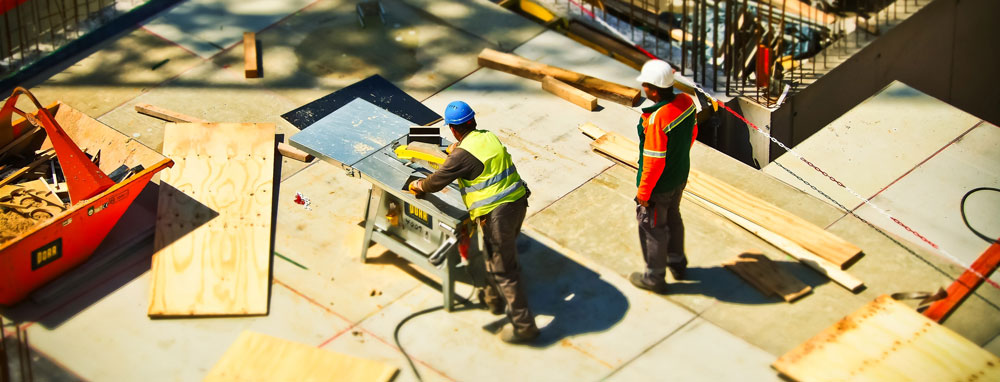 Two construction workers in safety vests and hard hats operate a circular saw on a concrete slab at a construction site.