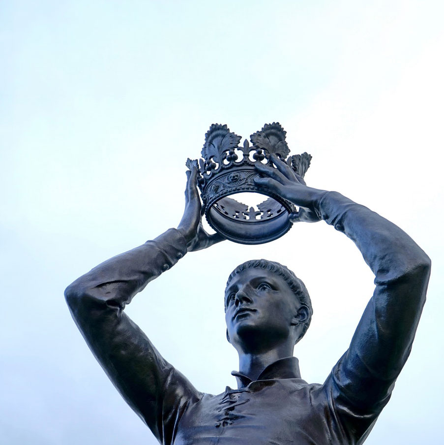 A dark bronze statue of a man holding a crown above his head.
