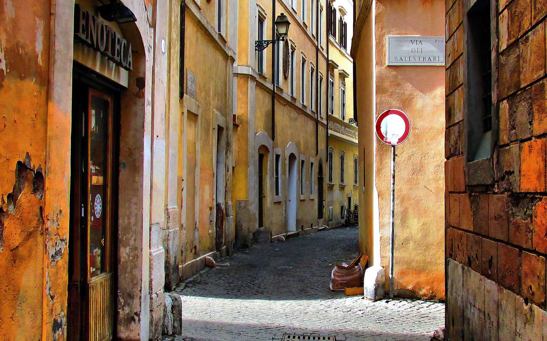 A narrow cobblestone street lined with yellow and orange buildings leads towards a sign reading “VIA DI LALESTERA”.