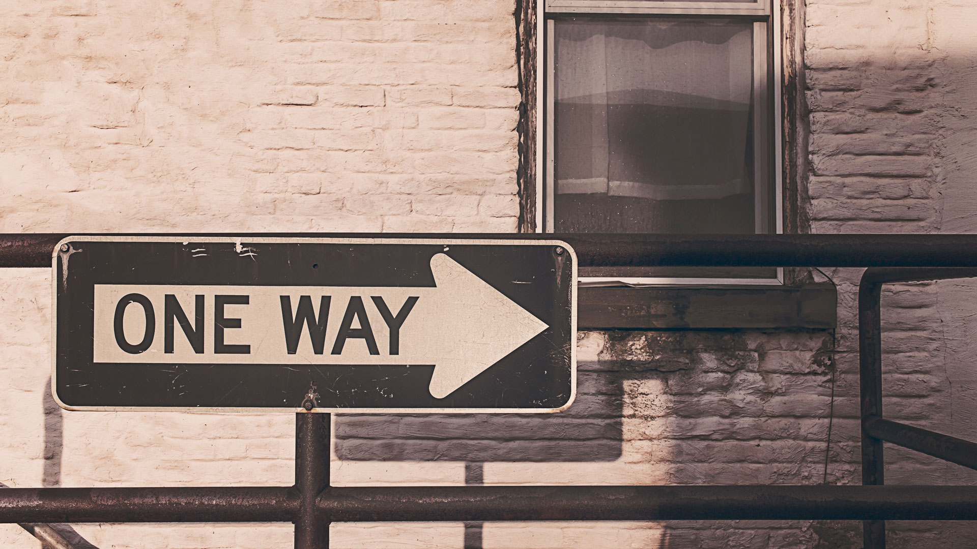 A black one-way sign with a white arrow points right is mounted on a metal railing in front of a white brick wall.