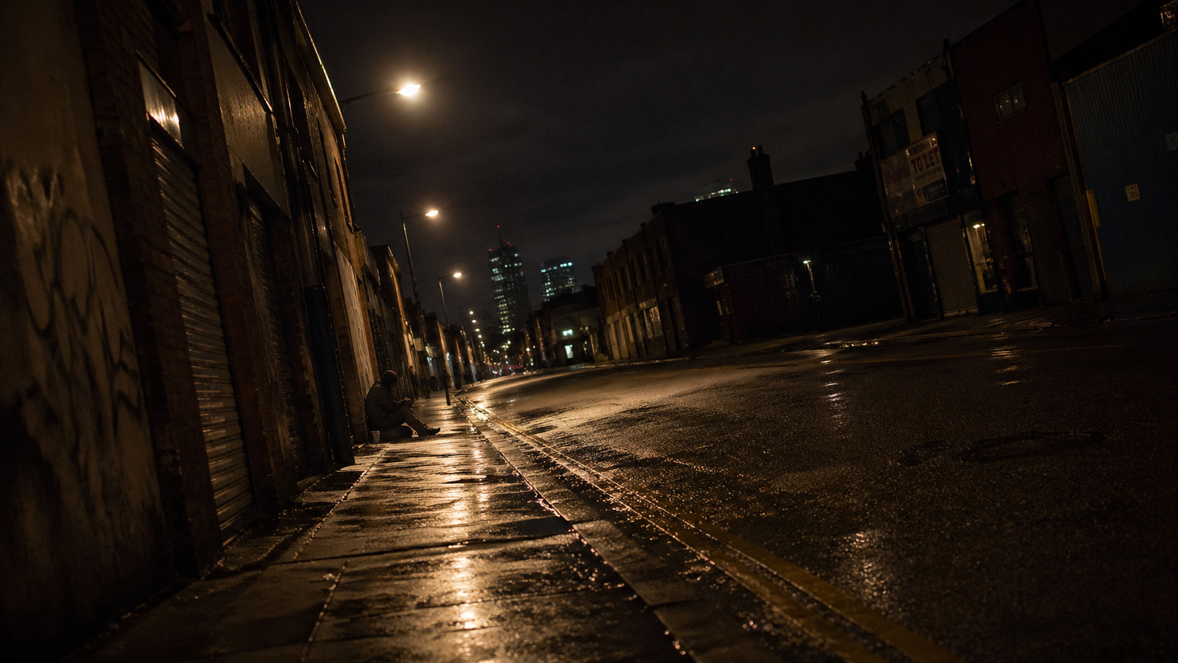 A wet, dark street at night with streetlights illuminating the wet pavement and buildings lining both sides. Graffiti is visible on the left wall, and a tall building with lit windows stands in the